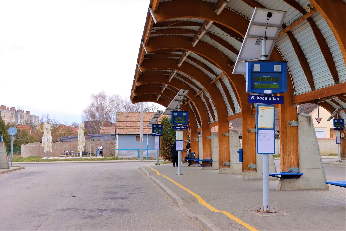A képen Szécsény autóbusz-állomás kocsiállásai láthatóak. A kocsiállásokat íves faszerkezet védi az esőtől és naptól, illetve napelemes utastájékoztató rendszerek kerültek kiépítésre, amiket a kép mutat.
