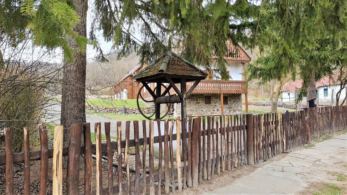 A képen Benczúrfalva rajziskolájának épülete látható. Előtérben kerekes kút, illetve fa kerítés látható, háttérben az erdő.