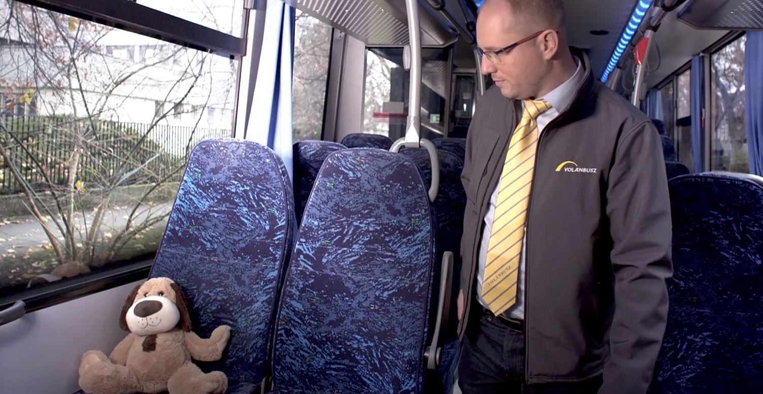A képen egy autóbusz belseje látható, az ülések között egy autóbuszvezetővel.