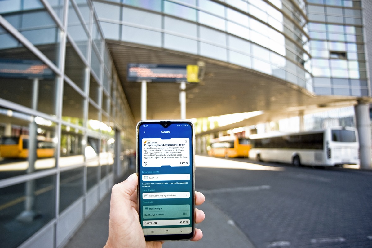 A képen egy telefonon megnyitott volánbusz applikáció látható, háttérben a népliget autóbuszállomás.